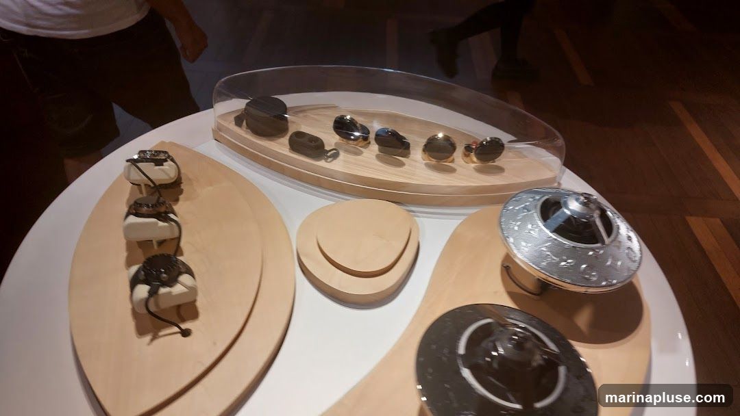 A display table showcases several luxury watches and other accessories. Three watches are presented on white stands on a wooden oval platform. In the background, a clear display case holds a collection of watches or similar items. Another ornate silver object is visible on the right side of the table. The setting appears to be a retail environment or exhibition.