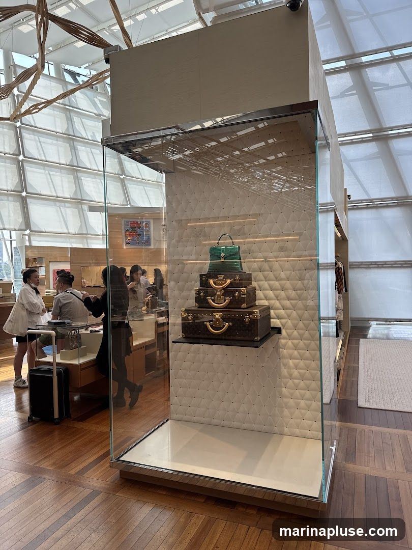 A display case showcases stacked Louis Vuitton luggage and a green handbag. In the background, customers interact with staff in a well-lit retail environment with wooden floors and a modern ceiling structure.