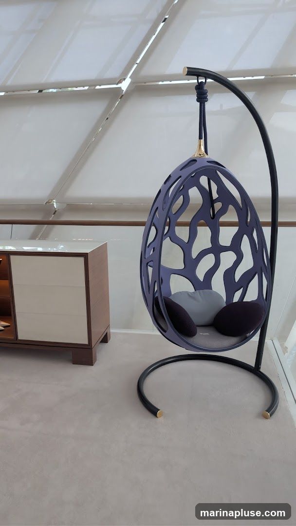 A modern, dark purple hanging egg chair with a decorative, organic-shaped exterior is shown. It is suspended from a black metal stand with gold accents. Inside the chair, there are three cushions: two dark purple and one light gray. To the left of the chair is a sleek, modern cabinet with a light wood frame and cream-colored drawers. The floor is carpeted in a neutral gray, and the ceiling features a translucent, paneled design.