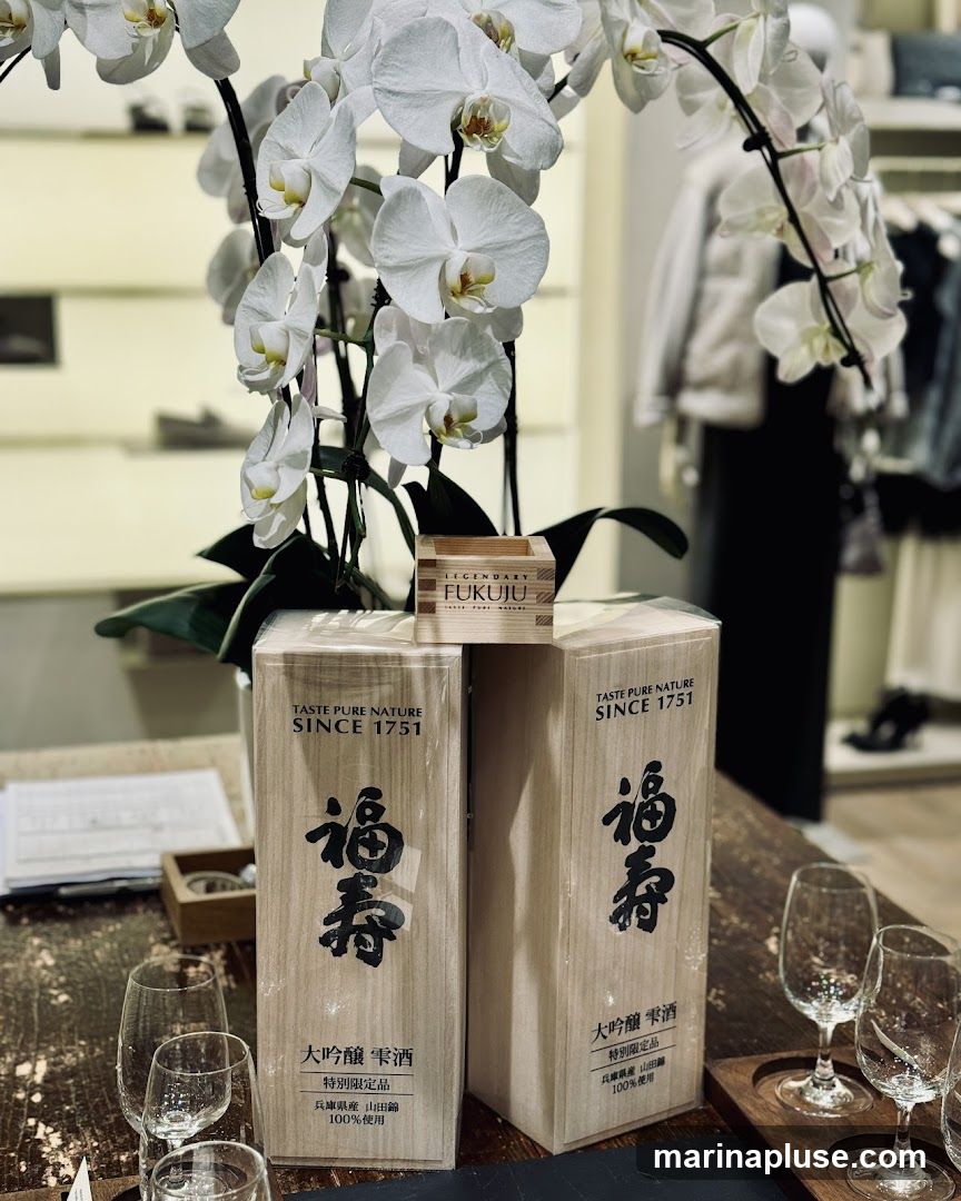 A display of Fukuju sake bottles, featuring white orchids and wine glasses. The wooden boxes have Japanese calligraphy and text indicating "Taste Pure Nature Since 1751" and "Daiginjo Shizuku Sake, Special Limited Edition, 100% Hyogo Prefecture Yamada Nishiki". A small wooden sake cup with "FUKUJU" branding is placed on top of the bottles. The background shows a blurred retail environment with clothing racks and shelves.