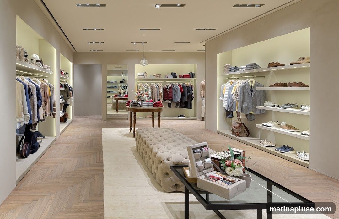 A well-lit, modern clothing store interior with herringbone wood flooring. Racks of men's clothing, including jackets, shirts, and sweaters, are displayed on shelves and hanging racks. Shoes are also displayed on shelves. In the foreground, a tufted ottoman and a glass-top table with a display box of accessories and flowers are visible. The store appears to be upscale and organized.