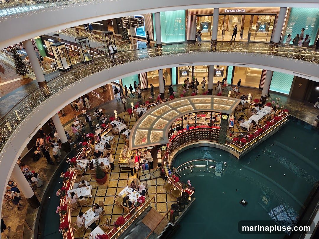 An aerial view of a multi-level indoor shopping mall featuring a canal with boats. The mall is decorated with festive lights. Several shops are visible on different levels, including Tiffany & Co. People are seen shopping and dining at tables set up around the canal.