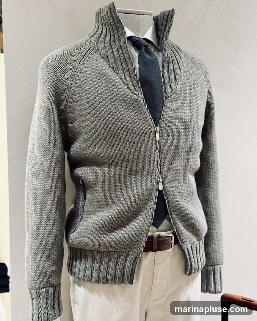 A mannequin is dressed in a grey knit zip-up cardigan over a white collared shirt and a dark grey tie. The outfit is completed with light-colored trousers and a brown belt. The background is a plain, textured wall.