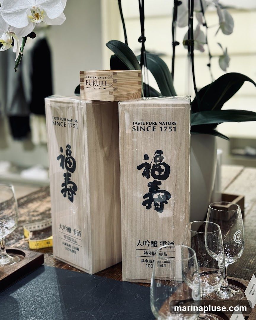 A display of Fukuju sake bottles and a wooden sake cup, with orchids and glassware in the foreground. The bottles and cup feature Japanese calligraphy and the text "Taste Pure Nature Since 1751".