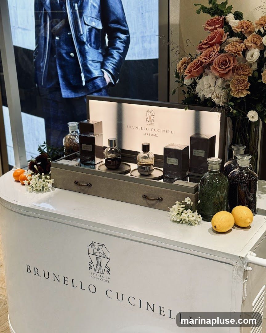 A display of Brunello Cucinelli perfumes is set up on a white counter. The display features several bottles of perfume, some in boxes, arranged on a grey tray. Behind the perfumes is a lit sign with the Brunello Cucinelli logo and the word "PARFUMS". To the right, a vase of flowers adds a decorative touch. The counter itself has the Brunello Cucinelli logo and name printed on it. A large image of a person in a denim jacket is visible in the background.
