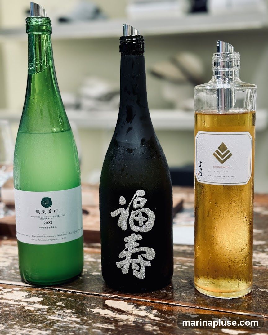 Three bottles of sake are displayed on a wooden surface. The bottle on the left is green and has a white label with Japanese text and "2023". The middle bottle is black with large, glittery silver Japanese characters. The bottle on the right is clear glass with a golden liquid and a white label that reads "KOZAEMON SINCE 1702 SAI-JIKOMI-KIJOSHU". All bottles have metal pour spouts. The background is blurred and appears to be a shelf with some items on it.
