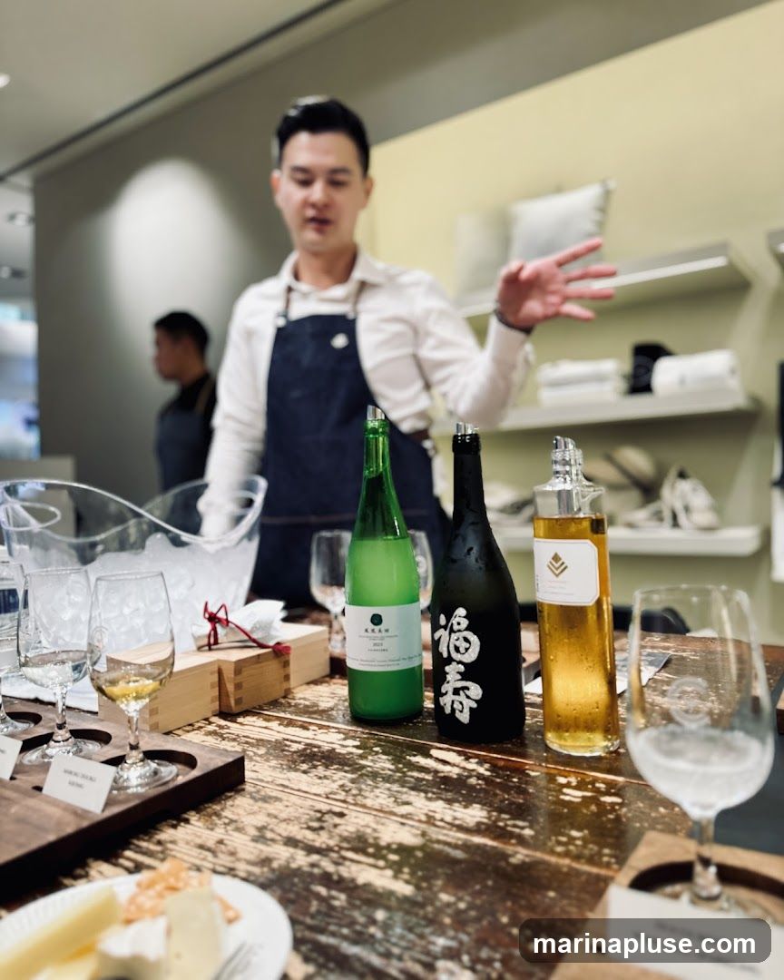 A man in an apron is gesturing while standing behind a table with three bottles of sake and several glasses. There is also a plate of cheese and crackers in the foreground. Shelves with items are visible in the background.