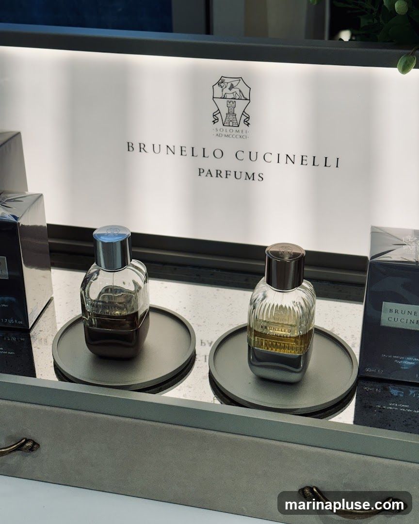 A display featuring two bottles of Brunello Cucinelli perfumes on a mirrored surface, with a backlit sign above that reads "BRUNELLO CUCINELLI PARFUMS" and the brand's crest. The bottles are on circular grey stands. There are also boxes of the perfume visible on either side.