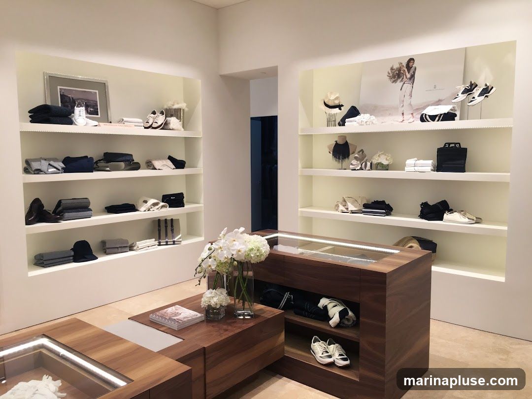 A well-lit retail space with shelves displaying folded clothing, shoes, hats, and accessories. A central wooden display counter features more merchandise. A large promotional image of a model is visible on the back wall.