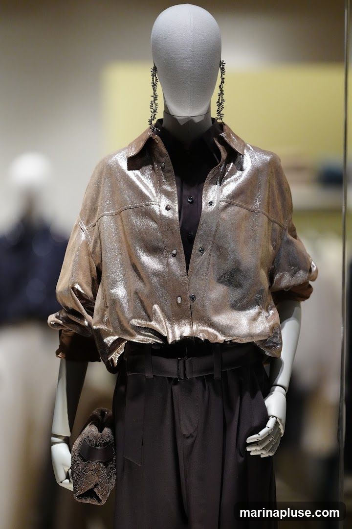 A mannequin is displayed wearing a shiny, metallic bronze-colored shirt over a dark collared shirt. The mannequin also wears dark, wide-legged trousers with a belt. Long, dangling earrings with a dark, intricate design are worn. A small, dark clutch bag with a lace-like pattern is held in the mannequin's left hand. The background is blurred, with hints of other mannequins and displays.