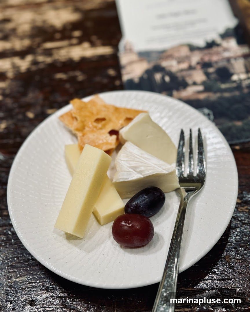 A white textured plate holds a selection of cheeses, crackers, and grapes. A fork rests on the side of the plate. In the background, a blurred menu or brochure displays a scenic image of a European town. The table surface is dark and appears to be made of wood.