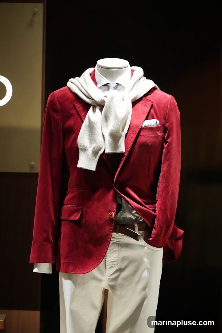 A mannequin is dressed in a red velvet blazer, a white dress shirt, a grey tie, and light-colored pants. A cream-colored scarf is draped around the mannequin's neck and over the blazer. A pocket square with a subtle pattern is visible in the blazer's breast pocket. The mannequin is displayed against a dark background with a hint of a white circular logo on the left side.
