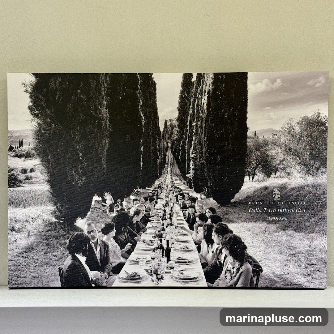 A black and white photograph depicts a long, formal outdoor dining table set for a large gathering. The table is laden with place settings, bottles of wine, and floral arrangements. People are seated on both sides of the table, engaged in conversation and dining. The scene is framed by tall cypress trees on either side, with a rolling landscape in the background. The Brunello Cucinelli logo and text "Dalla Terra tutto deriva SENOFANE" are visible on the right side of the image.