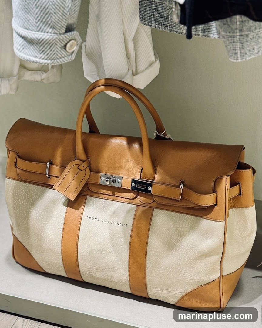 A close-up shot of a tan and cream leather duffle bag with the name BRUNELLO CUCINELLI embossed on the front. The bag has leather handles and trim, and a metal clasp closure. It is displayed on a shelf with clothing hanging above.