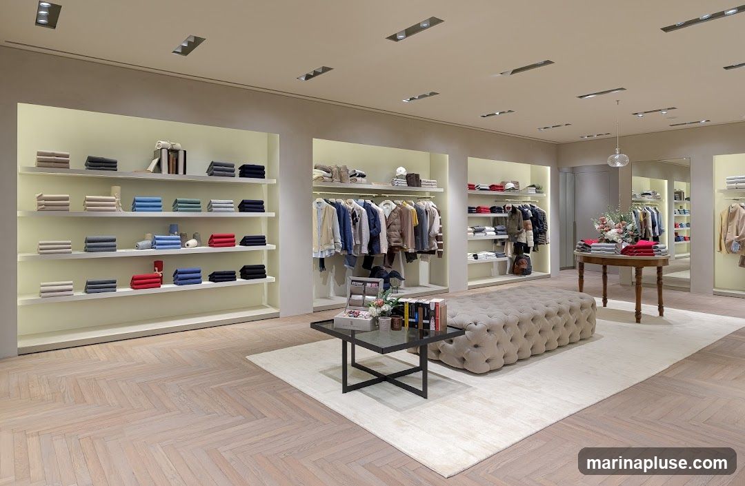 The image shows the interior of a well-lit, modern clothing store. On the left, shelves display neatly folded sweaters in various colors including beige, grey, blue, and red. In the center, racks of clothing, primarily jackets and shirts, are displayed. A large, tufted ottoman sits on a rug in the foreground. To the right, a round wooden table displays more folded clothing and a floral arrangement. Further back, more shelves with clothing and mirrors are visible. The flooring is light-colored wood laid in a herringbone pattern.