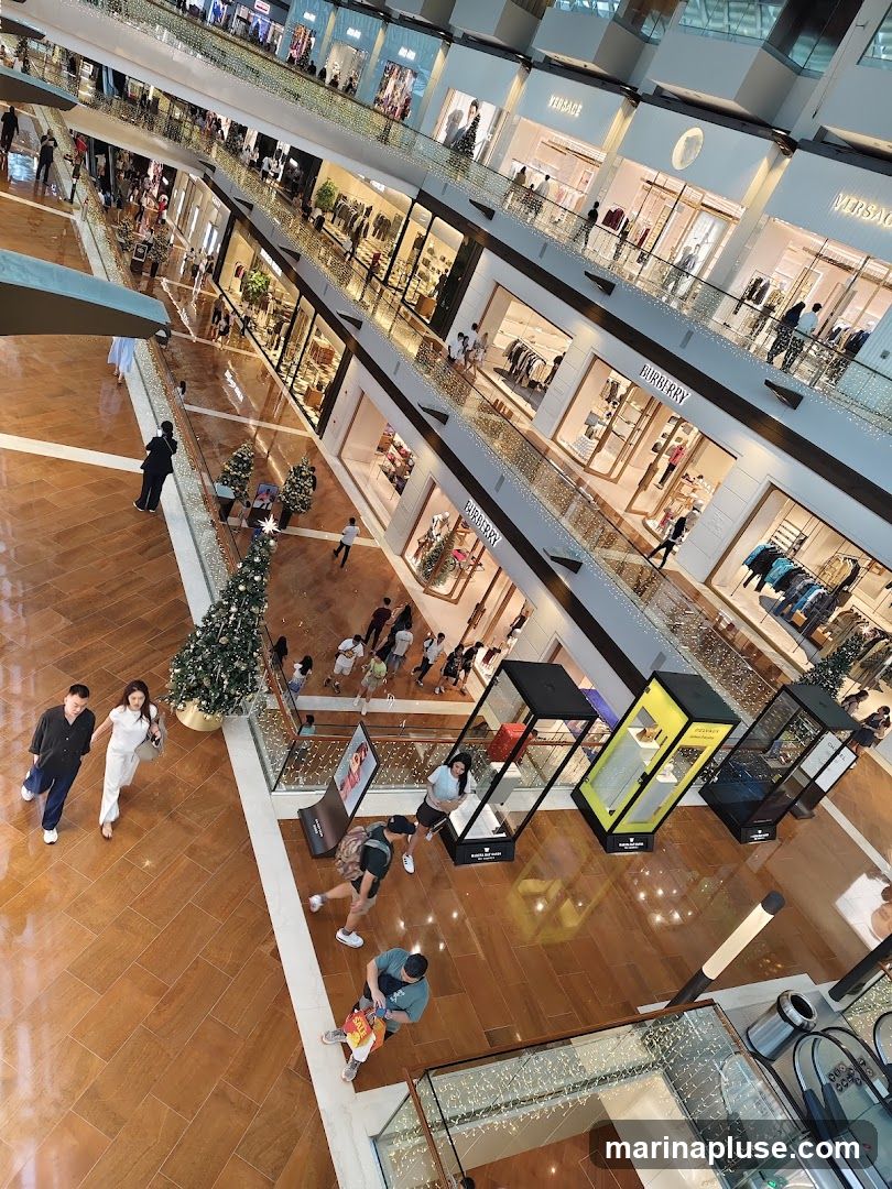 An overhead view of a multi-level shopping mall with numerous stores and shoppers. The stores visible include Versace and Burberry. Display cases with merchandise are set up in the open areas. Christmas trees are placed throughout the mall.