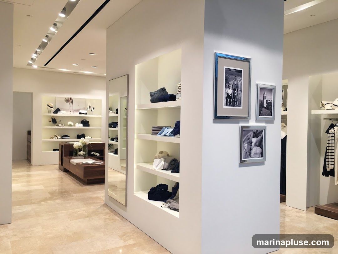 The interior of a modern, minimalist clothing store. Shelves display folded clothing, hats, and bags. A wooden display table with flowers and books is in the foreground. Three framed black and white photographs hang on a white pillar. Racks of clothing are visible in the background.