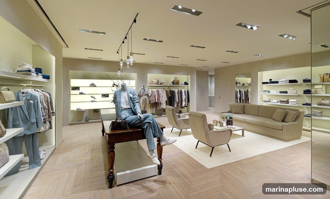 The image shows the interior of a well-lit, modern clothing store. A mannequin dressed in a light blue suit is prominently displayed on a dark wooden table. Racks of clothing, including jackets, shirts, and pants, are neatly arranged on shelves and hanging racks. Several pairs of shoes are displayed on illuminated shelves in the background. A comfortable-looking beige sofa and armchairs are arranged in a seating area, with a coffee table in front of them. The store features a clean, minimalist design with light-colored walls and wooden flooring laid in a herringbone pattern. Mirrors are strategically placed, reflecting the merchandise and enhancing the sense of space.