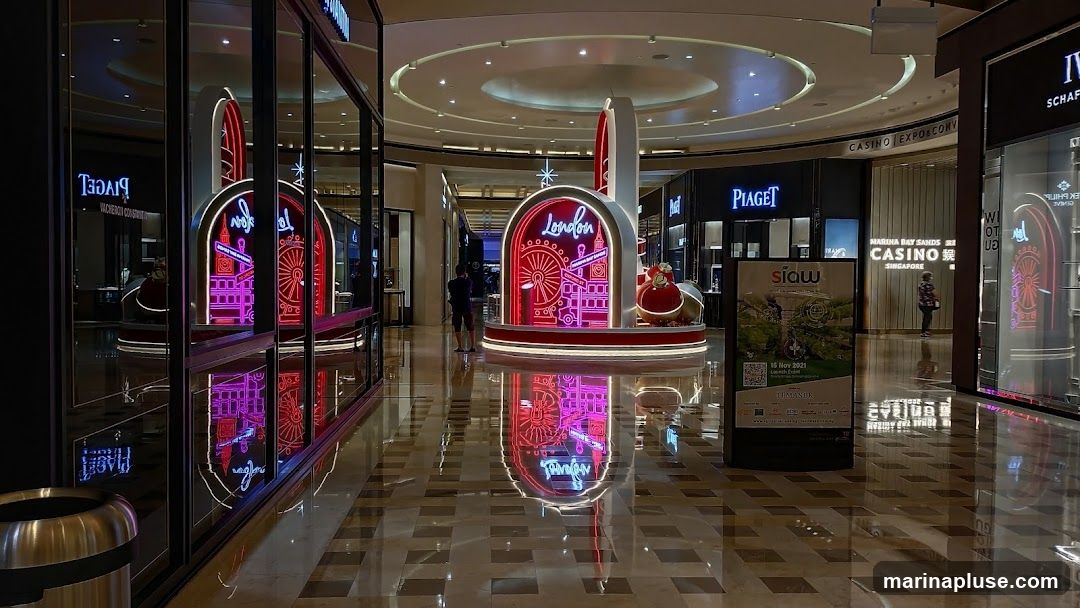 A wide shot of a mall interior featuring a large, brightly lit display with neon lights that spell out "London" and depict iconic London landmarks like the London Eye and Big Ben. The display is centrally located and reflects on the polished floor. Surrounding the display are various luxury storefronts, including Piaget and Vacheron Constantin, as well as a sign for a casino. A promotional poster for "siaw" is also visible.