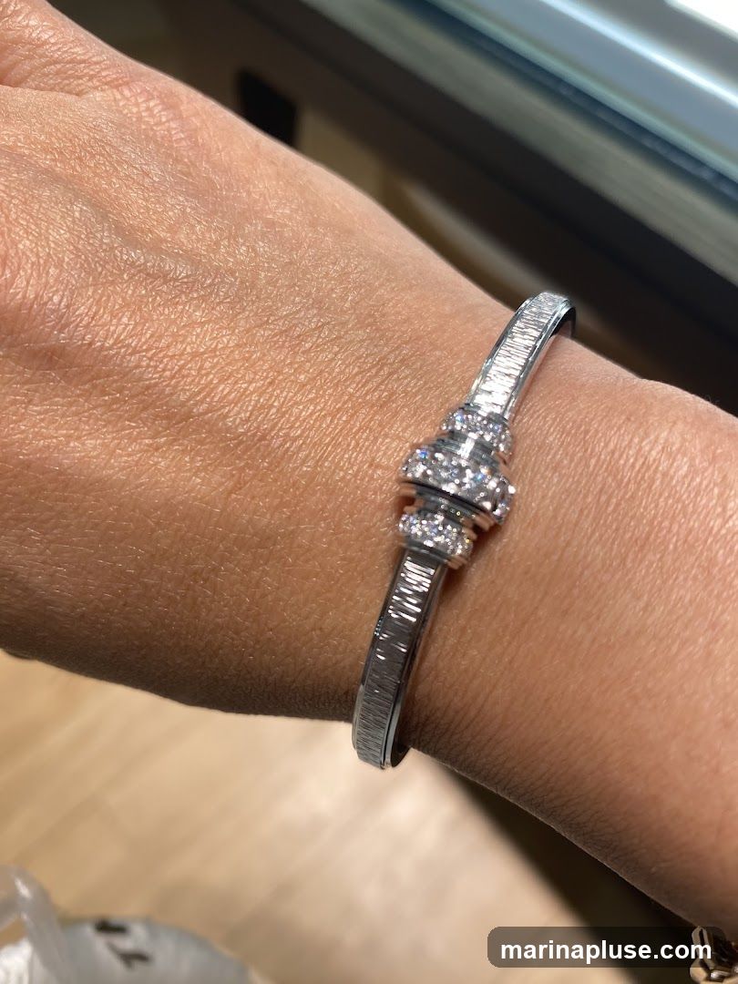 A close-up shot of a diamond and baguette bracelet on a person's wrist. The bracelet is made of white gold or platinum and features a central cluster of round diamonds surrounded by a band of baguette-cut diamonds. The band of the bracelet is also adorned with baguette-cut diamonds. The background is blurred, showing a wooden floor and a hint of a window.