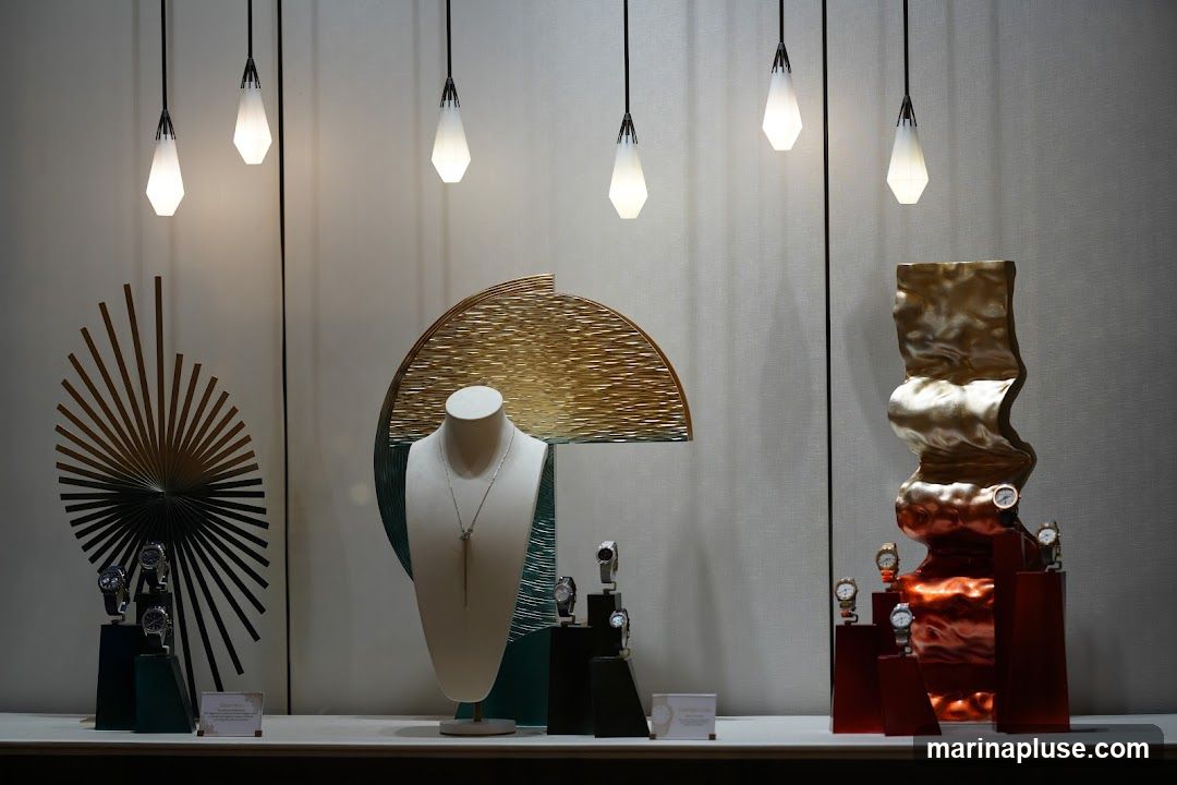 A well-lit display window showcases a variety of jewelry and watches. In the center, a mannequin bust wears a delicate silver necklace. To the left, a fan-like metallic sculpture is displayed alongside several watches on dark stands. To the right, a large, abstract, metallic sculpture in shades of gold and copper holds more watches. The background is a textured, neutral fabric, and several modern pendant lights hang from the ceiling. Small informational cards are placed at the base of some displays.