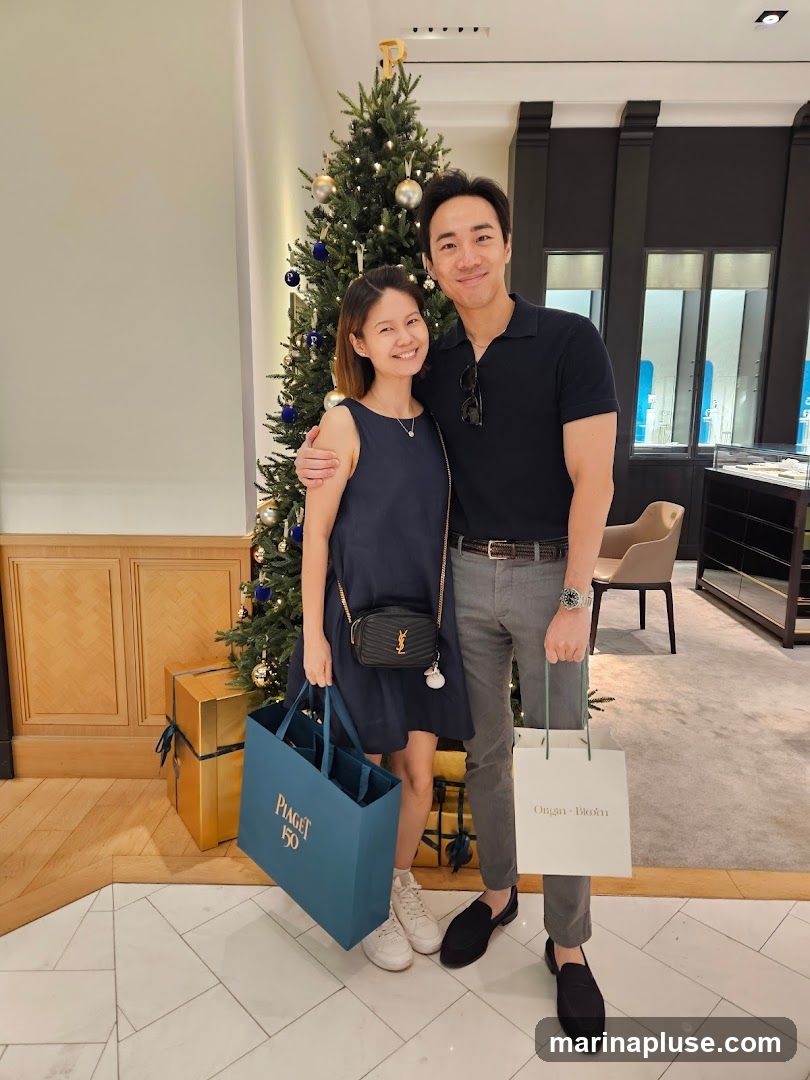 A couple smiles at the camera, standing in front of a decorated Christmas tree. The woman is wearing a dark blue dress and holding a teal shopping bag with "PIAGET 150" printed on it. The man is wearing a dark polo shirt and grey pants, holding a white shopping bag with "Ongan-Blohm" printed on it. They are in what appears to be a retail store, with display cases and furniture visible in the background.