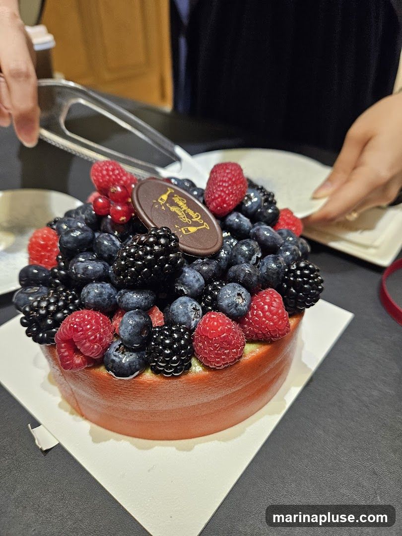 A round cake topped with a generous assortment of fresh berries, including blueberries, raspberries, and blackberries. A small oval chocolate plaque with gold lettering and an image of champagne flutes is placed on top of the berries. The cake has a reddish-orange exterior and appears to be on a white serving plate. Tongs are being used to serve the cake.