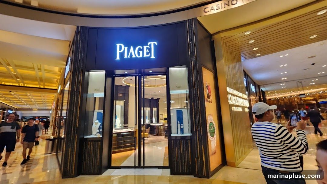 The exterior of a Piaget jewelry and watch store. The store has a dark facade with the illuminated Piaget logo above the entrance. The entrance features glass doors and display windows showcasing jewelry and watches. A large advertisement for a watch is visible on the right side of the storefront. People are walking by in the background.