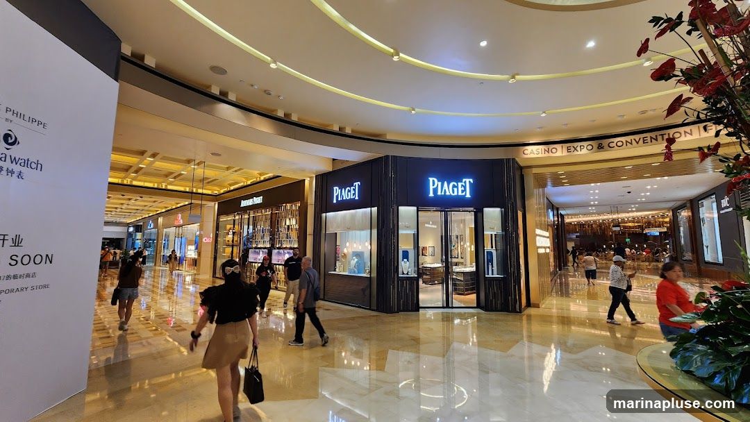 A wide shot of a brightly lit indoor mall with several luxury watch stores. The Piaget store is prominently featured in the center, with its name displayed in large illuminated letters. To the left, an Audemars Piguet store is visible. A large white banner on the far left advertises a store opening soon. People are walking through the mall, and the polished floors reflect the overhead lighting. In the background, signage indicates "CASINO | EXPO & CONVENTION."