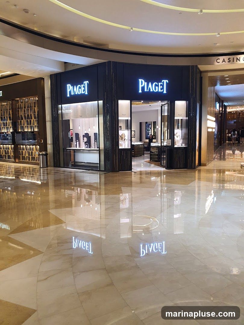 A well-lit Piaget store with its name prominently displayed in large, illuminated letters above the entrance. The storefront features large glass windows showcasing displays of watches and jewelry. The interior is visible through the entrance, revealing more display cases and a person standing inside. The store is located in a modern, upscale shopping mall with polished marble floors that reflect the store's signage.