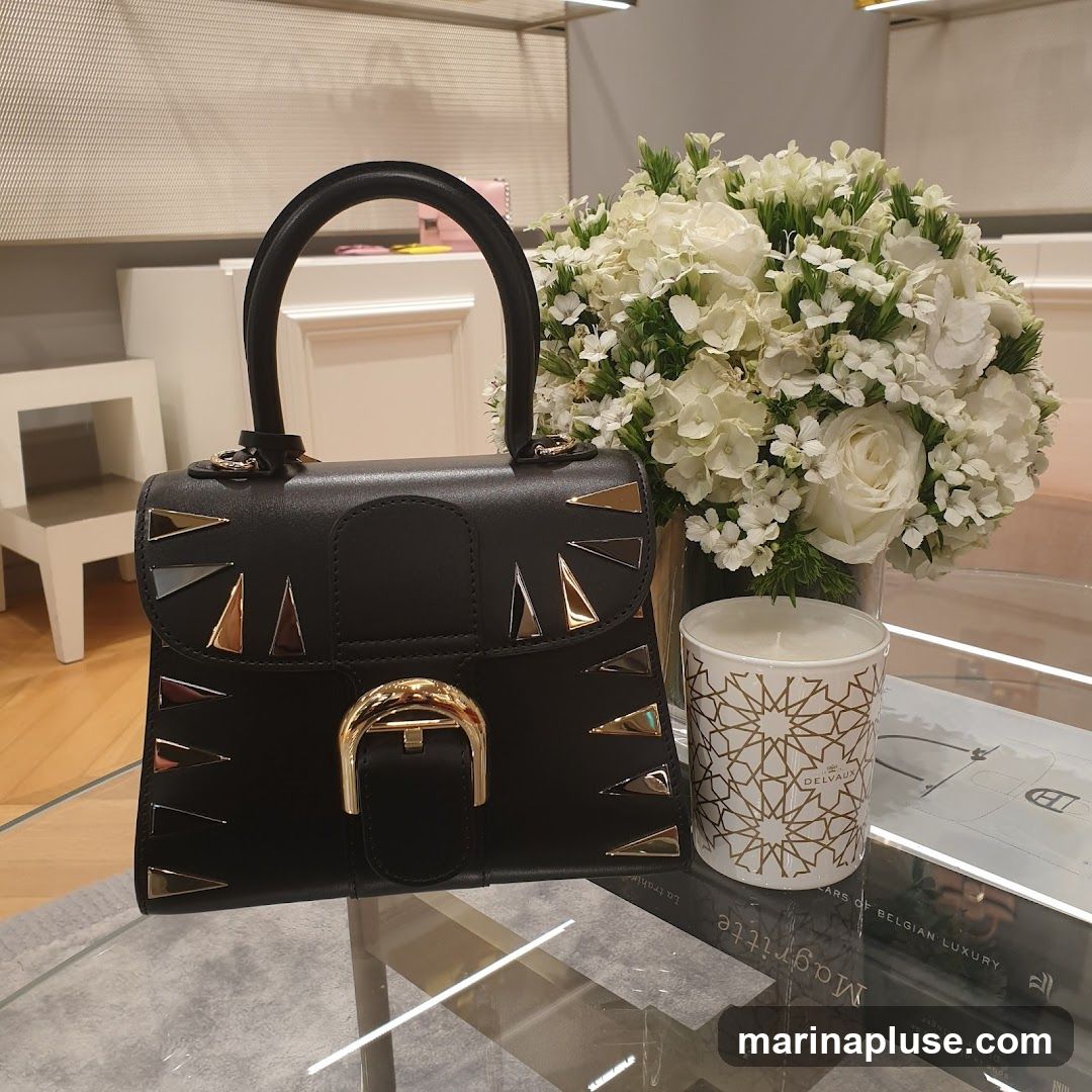 A black leather handbag with gold and silver triangular accents is displayed on a glass table. Next to it is a bouquet of white flowers and a white candle with a gold geometric pattern. A book titled "Magritte" is visible beneath the items. The brand name "DELVAUX" is visible on the candle.