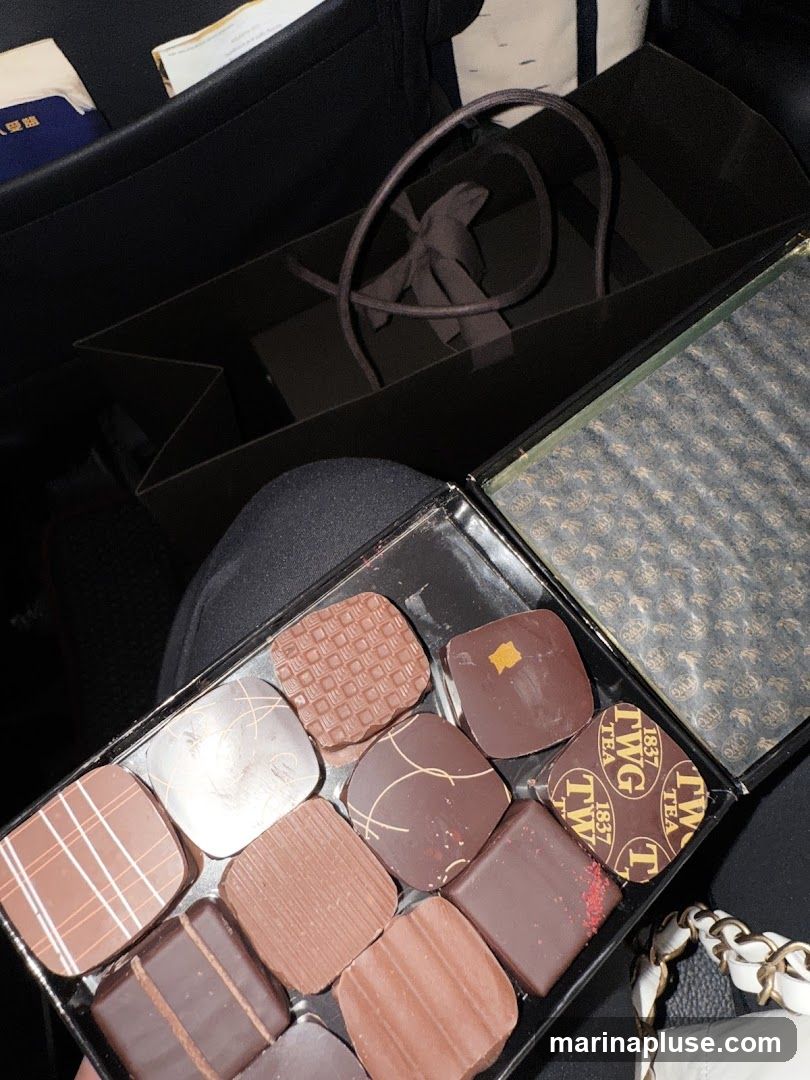 A box of assorted chocolates with various designs and toppings. Some chocolates have intricate patterns, while others are plain or decorated with edible gold leaf or sprinkles. The box is open, revealing the chocolates arranged in compartments. A shopping bag and other items are visible in the background. The lid of the box has a repeating pattern with the text "TWG 1837 TEA" visible on some of the chocolates.