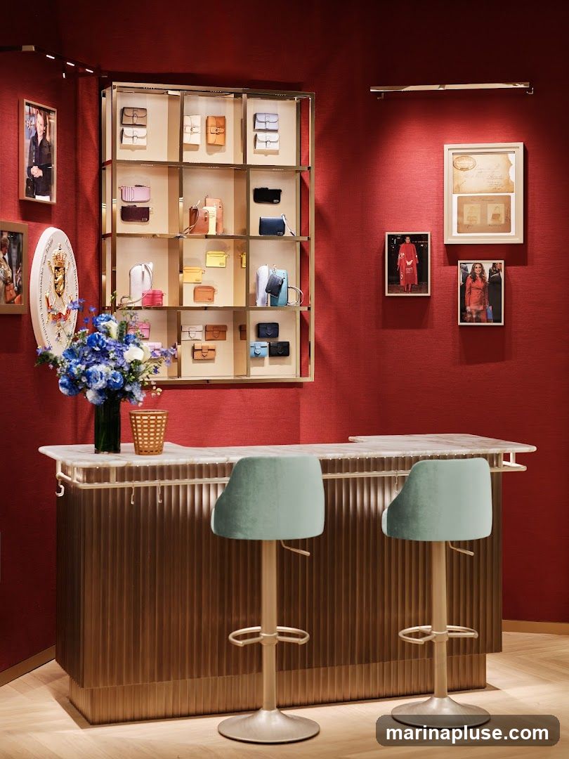 A retail interior featuring a display of various handbags on shelves against a textured red wall. In the foreground, a marble-topped bar with a fluted gold base is set up with two light blue velvet bar stools. Artwork and framed photographs adorn the walls.