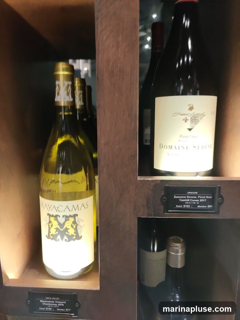 Several bottles of wine are displayed on shelves. One bottle in the foreground on the left is labeled "MAYACAMAS Napa Valley Mayacamas Vineyard Chardonnay 2018". To the right, a bottle labeled "DOMAINE SERENE Oregon Domaine Serene, Pinot Noir Yamhill Cuvee 2017" is visible with pricing information below. Other bottles are partially visible in the background and on lower shelves.