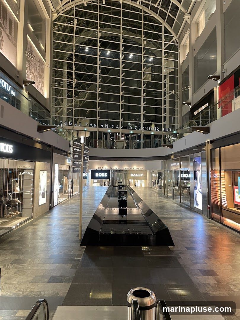 A view down a wide, modern shopping mall interior with a reflective water feature in the center. Shops with illuminated displays line both sides of the walkway. The architecture features a large, arched glass ceiling.