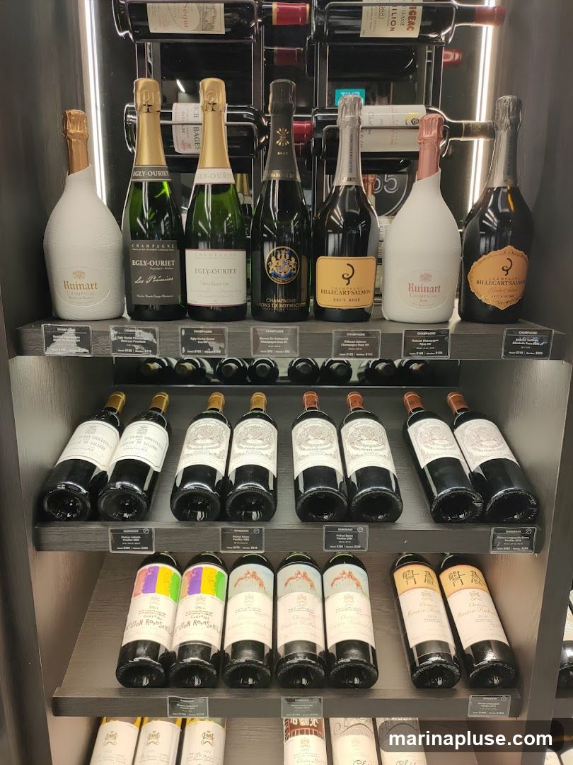 A display of various bottles of wine and champagne on shelves. The top shelf shows red wine bottles in racks. The second shelf features several bottles of champagne, including Ruinart, Egly-Ouriet, and Billecart-Salmon. The third shelf displays red wine bottles, possibly Bordeaux. The fourth shelf shows more red wine bottles, some with colorful labels. The bottom shelf is partially visible with more wine bottles. Small price tags are visible below some of the bottles.