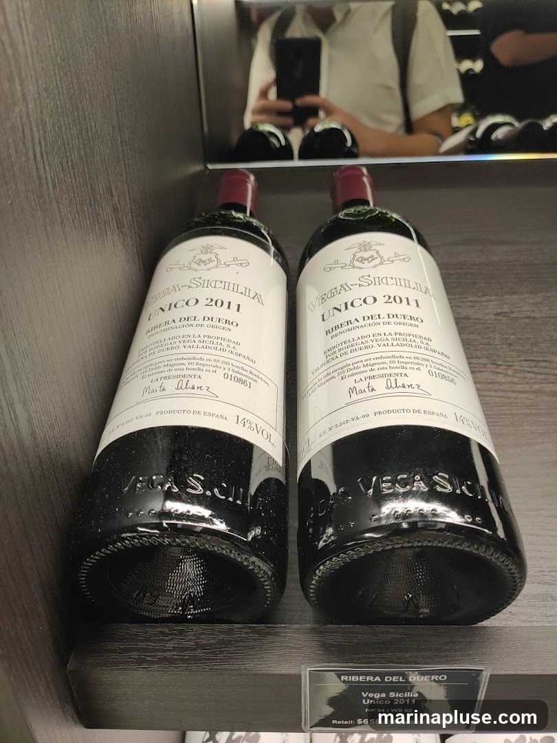 Two bottles of Vega Sicilia Unico 2011 wine are displayed on a shelf. The labels indicate they are from the Ribera del Duero region of Spain and have a 14% alcohol by volume. A price tag below shows the retail price as $658 and the member price as $618. A reflection in the background shows a person holding a phone and taking a picture.
