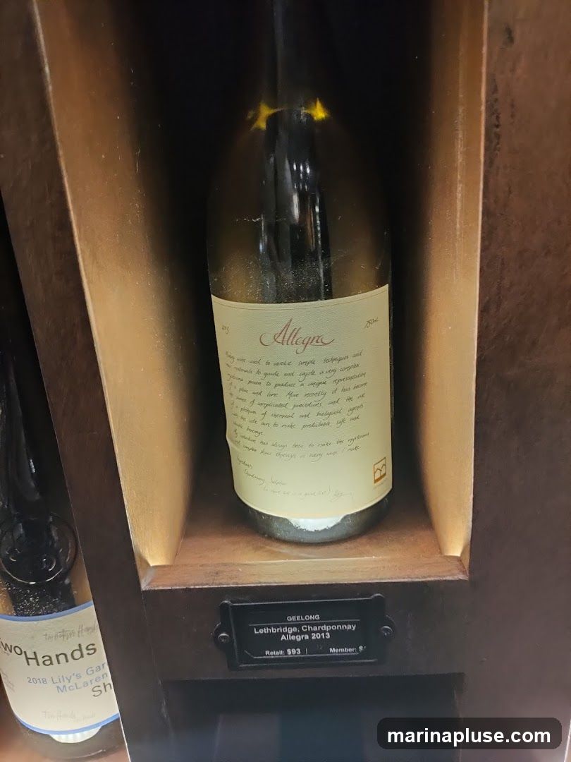 A close-up view of a wine bottle with a descriptive label, displayed in a wooden shelf with interior lighting. Another wine bottle is partially visible to the left. A small metal plaque below the featured bottle reads "GEELONG Lethbridge, Chardponnay Allegra 2013 Retail: $93 Member: $".
