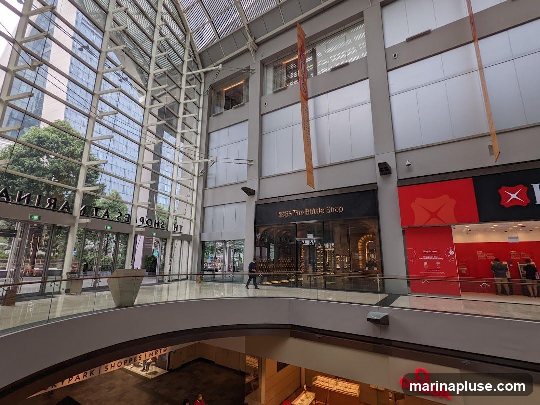 An interior view of a modern shopping mall with a glass atrium and multiple levels. On the upper level, storefronts are visible, including '1955 The Bottle Shop' and a red-branded bank. A person is walking on the upper walkway. Below, signage for 'SKYPARK SHOPPES MRT' and an 'OMEGA' store are visible.