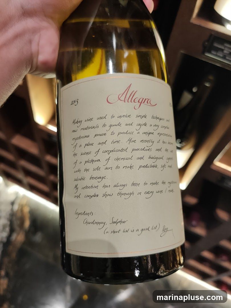 A close-up shot of a wine bottle with a label that reads "Allegra" and the year "2013". The label contains a description of the winemaking philosophy and lists ingredients as Chardonnay and Sulphur. The bottle is held by a hand and appears to be in a wine cellar or display.