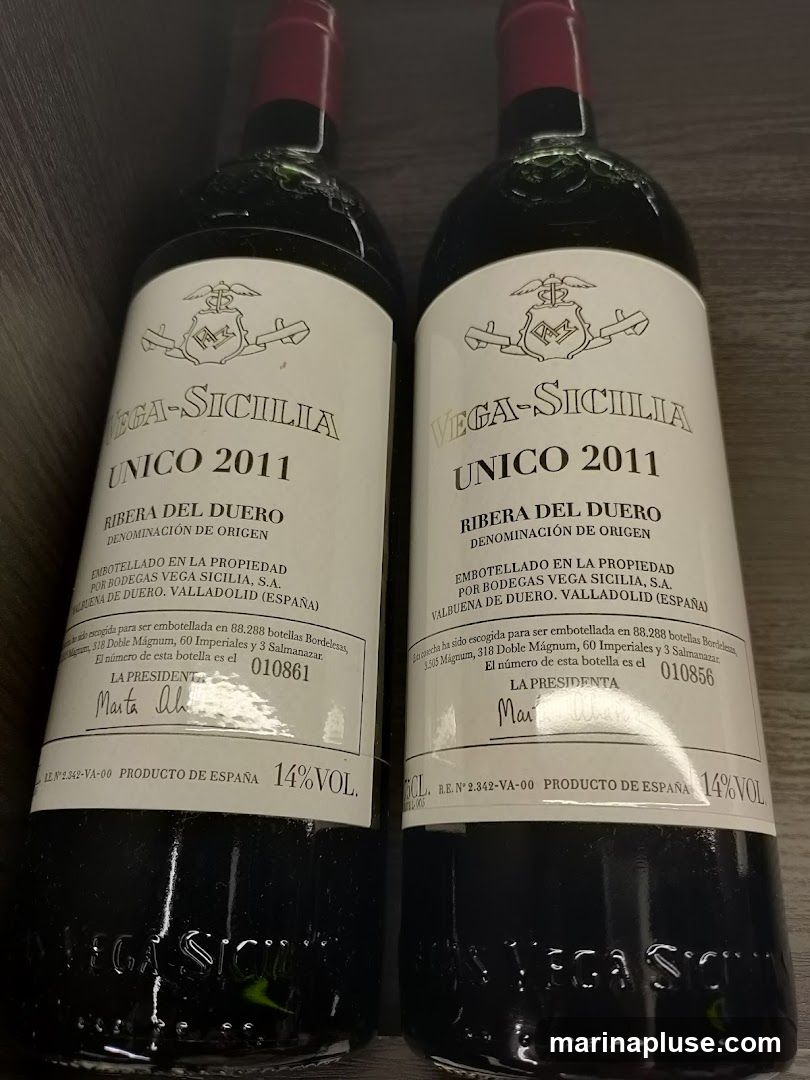 Two bottles of Vega-Sicilia Unico 2011 wine are displayed. The labels indicate they are from Ribera del Duero, Spain, bottled by Bodegas Vega Sicilia, S.A. in Valbuena de Duero, Valladolid. The text also mentions the total number of bottles produced and the specific bottle number for each. The wine has a 14% alcohol by volume.