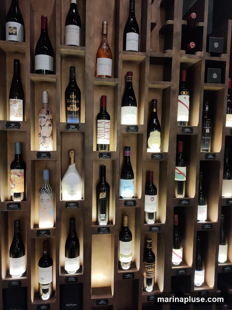 A wall display of numerous wine bottles arranged on shelves. The bottles vary in shape, color, and label design. Some bottles are illuminated from below, highlighting their contents and labels. The display appears to be in a wine shop or a similar establishment.