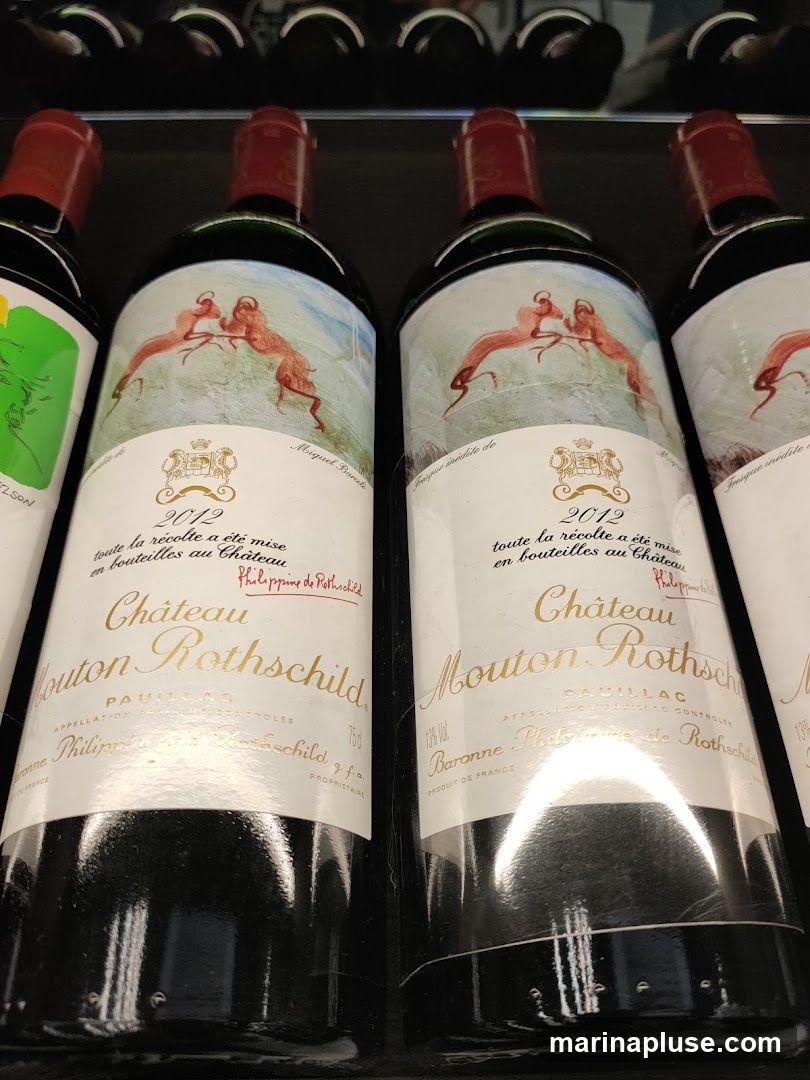 Several bottles of Chateau Mouton Rothschild wine are displayed in a rack. The labels feature artwork of two deer-like animals, the year 2012, and the text "Château Mouton Rothschild".