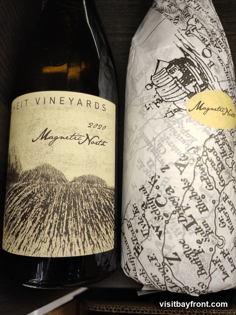 Two bottles of wine are shown. The bottle on the left has a label that reads "HEIT VINEYARDS 2020 Magnetic North" with an illustration of hills or fields. The bottle on the right is wrapped in paper with a map-like pattern and has a yellow oval sticker that says "Magnetic North" in cursive.