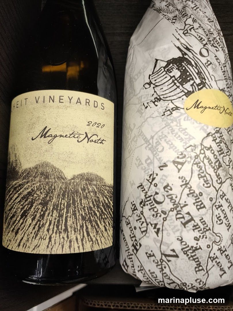 Two bottles of wine are shown. The bottle on the left has a label that reads "HEIT VINEYARDS 2020 Magnetic North" with an illustration of hills or fields. The bottle on the right is wrapped in paper with a map-like pattern and has a yellow oval sticker that says "Magnetic North" in cursive.