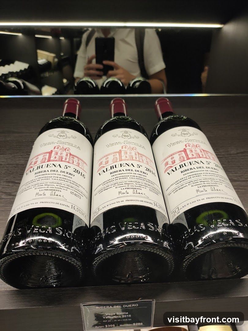 Three bottles of Vega Sicilia Valbuena 5° 2016 wine are displayed on a shelf. The labels are red and white, featuring an illustration of a building and text in Spanish and English. A price tag below the bottles indicates retail and member prices. In the reflection on the shelf above, a person is visible taking a photo with their phone.