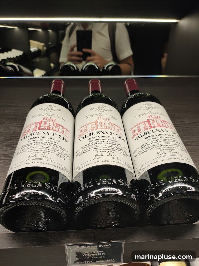 Three bottles of Vega Sicilia Valbuena 5° 2016 wine are displayed on a shelf. The labels are red and white, featuring an illustration of a building and text in Spanish and English. A price tag below the bottles indicates retail and member prices. In the reflection on the shelf above, a person is visible taking a photo with their phone.