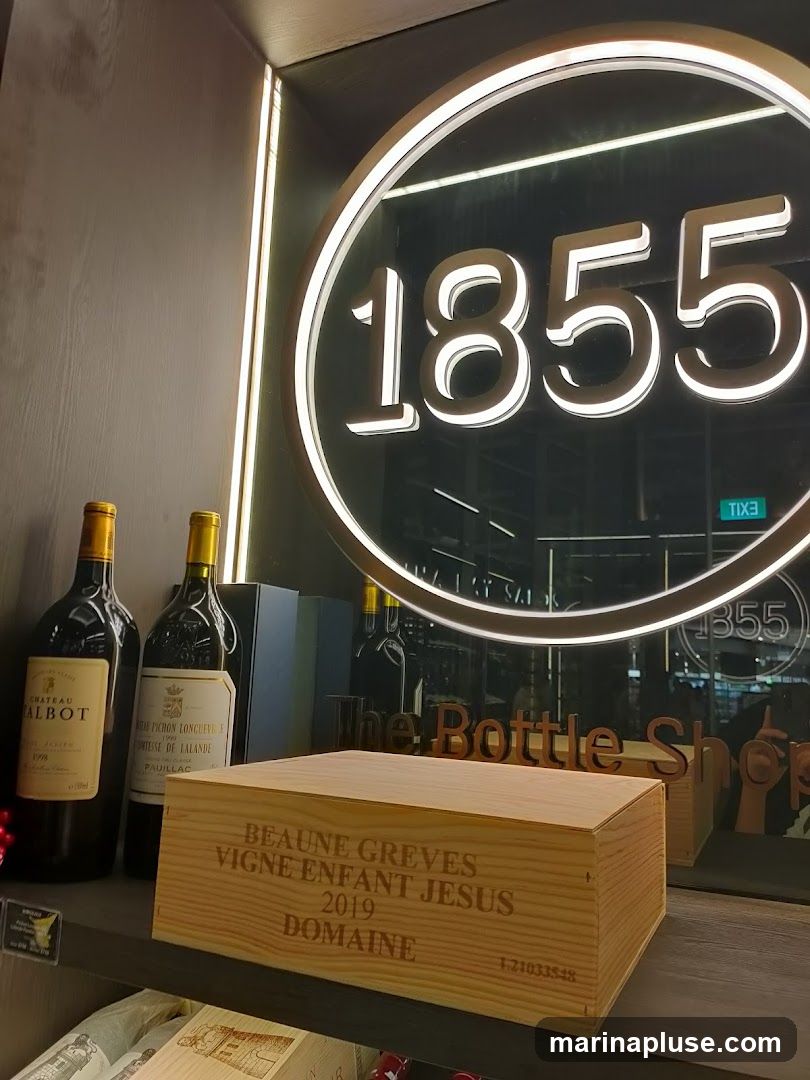 A wooden wine box with text 'BEAUNE GREVES VIGNE ENFANT JESUS 2019 DOMAINE' is displayed on a shelf in front of two bottles of wine. Behind the wine box, a large circular sign with the number '1855' illuminated in white is visible. The reflection in the glass behind the sign shows the words 'The Bottle Shop'.