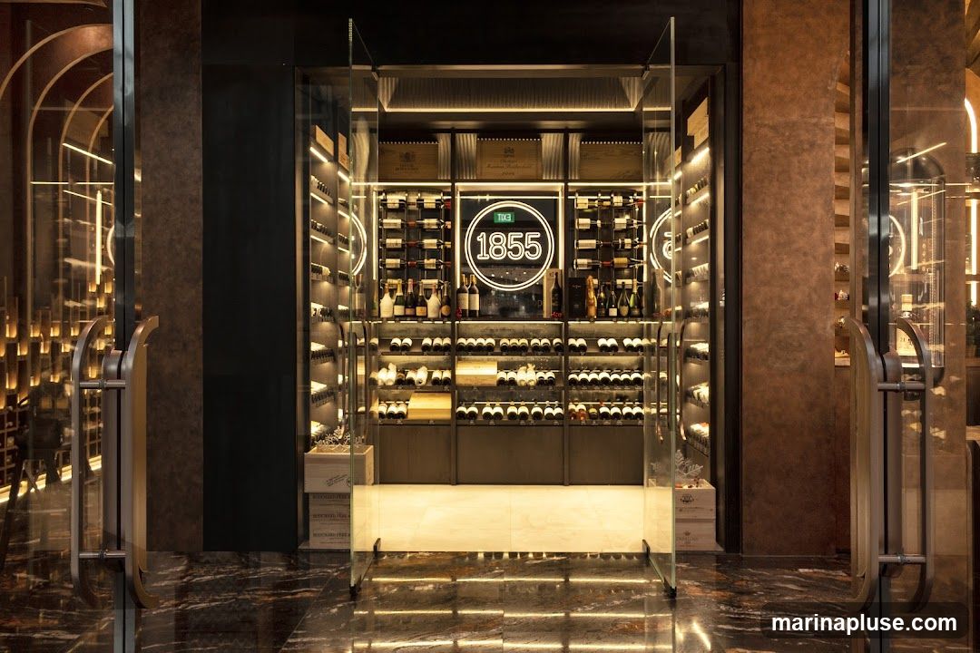 A well-lit wine cellar with rows of wine bottles displayed on shelves. A neon sign with the number "1855" is visible in the center. The entrance is framed by glass doors and metallic handles. The floor is made of polished marble with reflective lighting.