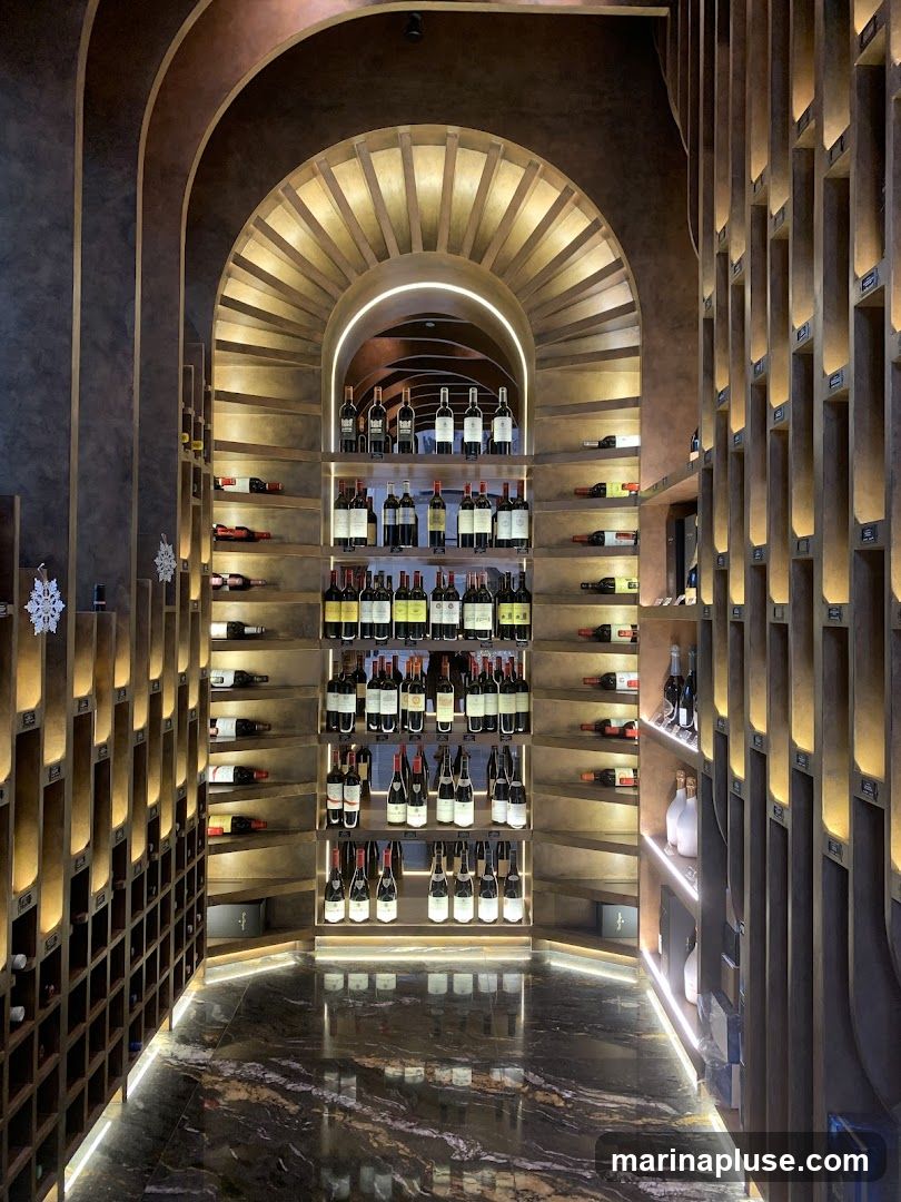 A well-lit wine cellar with extensive shelving filled with numerous bottles of wine. The shelves are arranged in a semi-circular fashion, with additional vertical shelving units on the sides. The floor is made of polished marble, reflecting the ambient lighting.